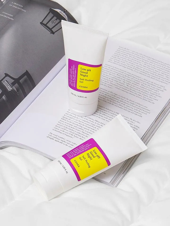 COSRX Low pH Good Night Soft Peeling Gel tubes on open magazine and white bedding, Korean skincare