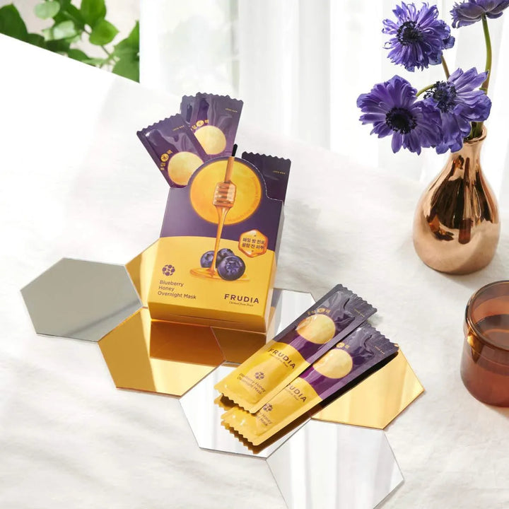 Frudia Blueberry Honey Overnight Mask sachets on a mirrored surface with purple flowers, Korean skincare Me Time Groningen