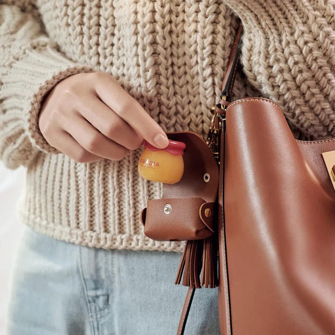 Hand holding Frudia Pomegranate Honey 3-in-1 lip balm near brown leather bag, Korean skincare
