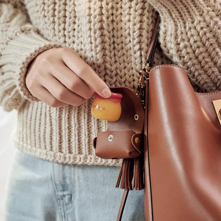 Hand holding Frudia Pomegranate Honey 3-in-1 lip balm near brown leather bag, Korean skincare