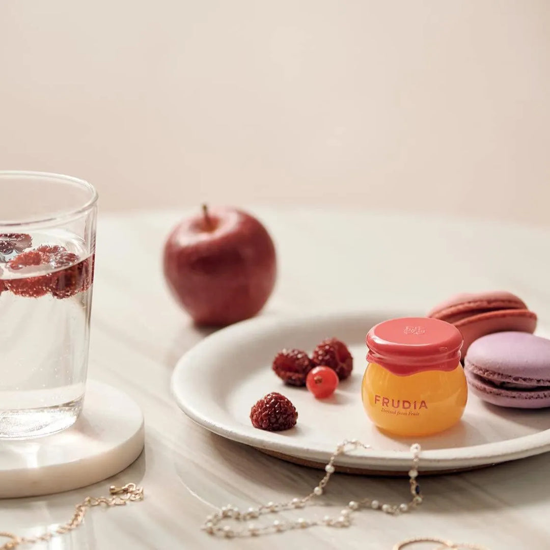 Frudia Pomegranate Honey 3-in-1 Lip Balm on a plate with berries, macarons, and apple, Korean skincare product at Me Time Groningen.
