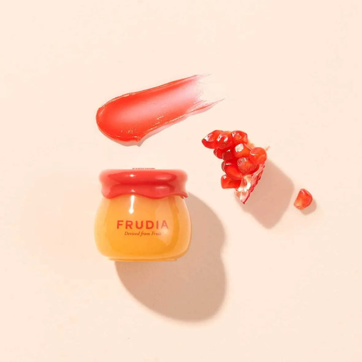 Frudia pomegranate honey 3-in-1 lip balm in jar, swatch, and pomegranate seeds on light background